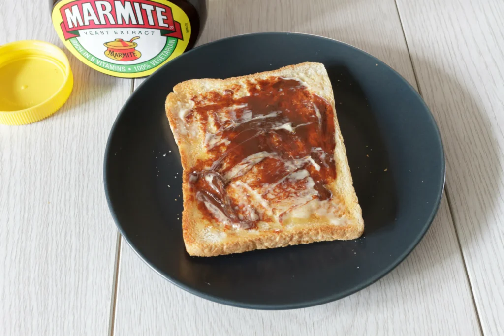 Тост с тонким слоем мармайта (Marmite) на тёмной тарелке, рядом банка пасты